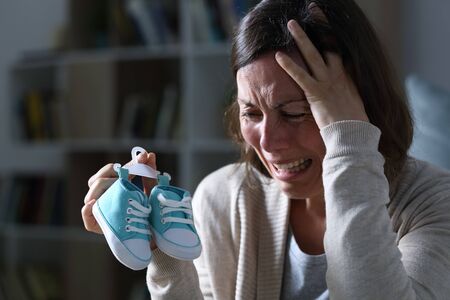 Sad mother crying missing her daughter after miscarriage holding new baby shoes at night at home