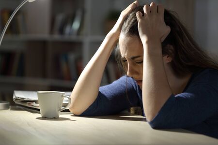 Sad woman complaining at night grabbing head sitting alone at home