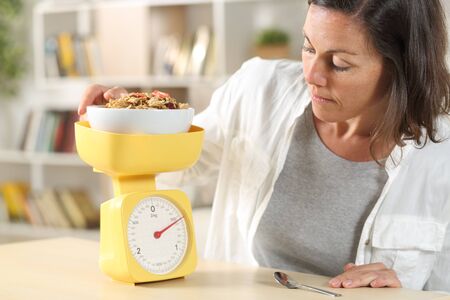 Adult woman dieting measuring food with scale weighting cereal bowl at homeの写真素材