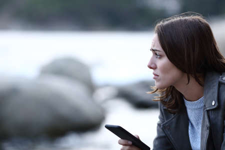 Sad woman looking away holding smart phone alone on the beach