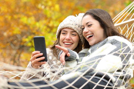 Two happy friends checking smart phone in a hammock in autumn season in a forestの写真素材