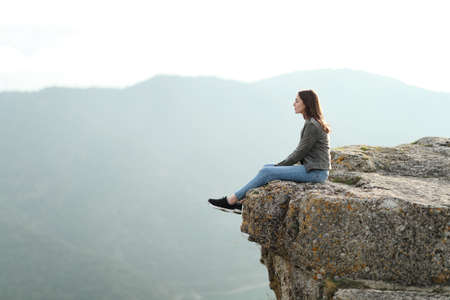 Full body profile of a pensive woman sitting contemplating views on the top of a cliff in the mountainの写真素材