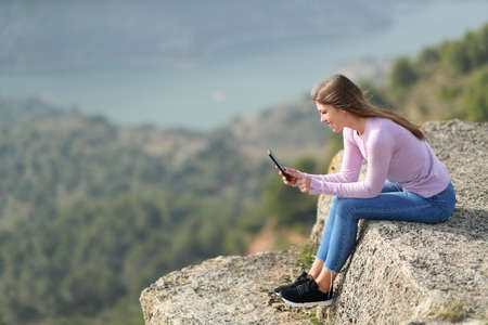 Full bofy side view portrait of a happy teen checking smart phone in a cliff