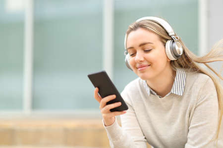 Satisfied teen listening to music on smart phone sitting in the street