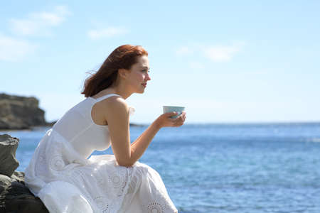 Profile of a woman in white dress drinking coffee contemplating ocean on the beach