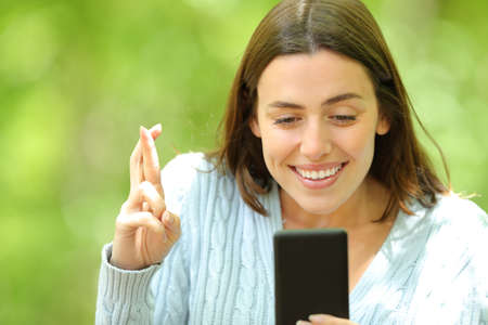 Hopeful woman crossing fingers checking smart phone in a green parkの写真素材