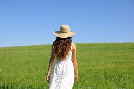 Back view portrait of a woman with white dress and pamela hat walking in green wheat field