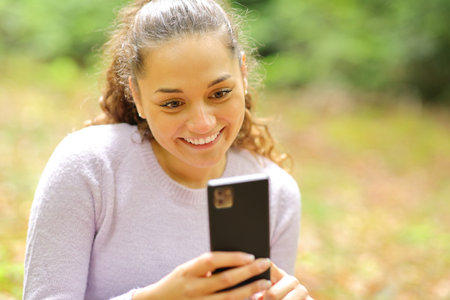 Happy woman sitting in a forest or park using smart phoneの写真素材
