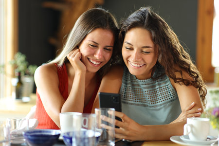 Front view of two happy friends checking smart pone in a restaurant