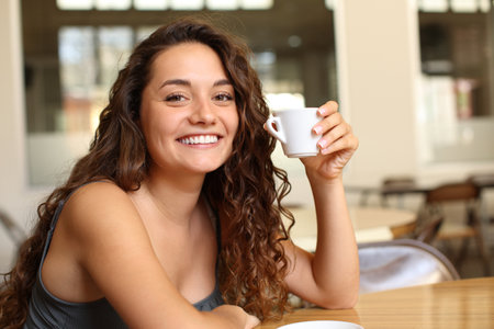 Happy woman holding coffee mug looking at you sitting in a bar and smilingの写真素材