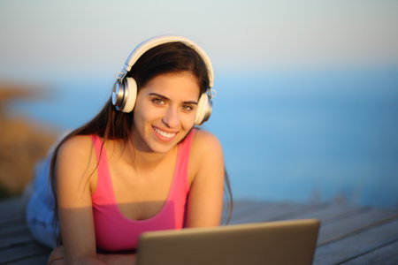 Happy woman with headphone and laptop posing looking at camera on the beachの写真素材