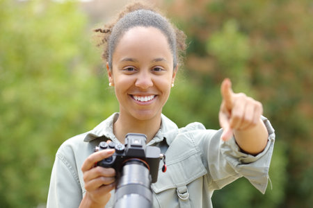 Front view portrait of a black photographer pointing at camera holding mirrorless camera in a parkの写真素材
