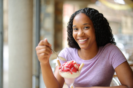 Happy black woman eating dessert in a restaurant terrace looking at cameraの写真素材