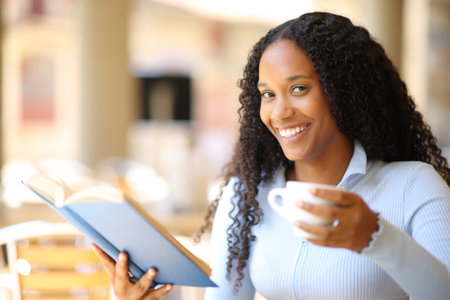 Happy black woman reading a paper book drinking coffee in a bar terrace