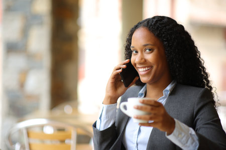 Happy black executive drinking and talking on phone looking at you in a coffee shop terraceの写真素材