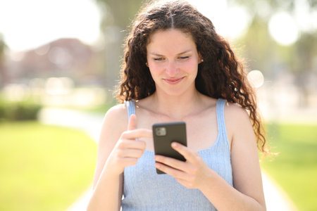 Front view portrait of a happy woman with curly hair using phone walking in a parkの写真素材