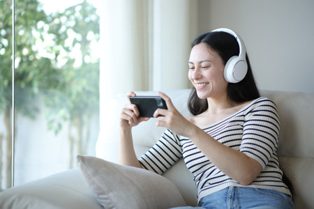 Happy asian woman with headphone watching media on phone sitting on a sofa at homeの写真素材