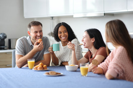 Happy interracial friends at breakfast talking eating and drinking in the kitchen at homeの写真素材