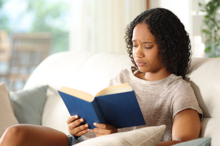 Worried black woman reading a paper book sitting on a sofa at homeの写真素材