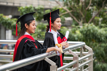 Two smiling female Asian students in graduation gown holding the diplomas with eyes looking forward