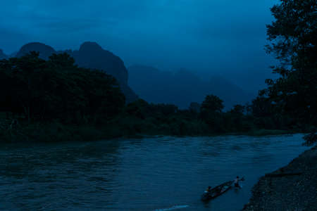 Two men in a small boat finish crossing a river at dusk in Vang Vieng, Laos, with mountains in the...
