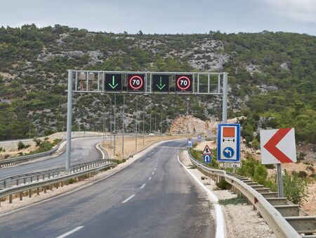 Asphalt road with many road signs and metal arch with direction arrows and speed limit signs.