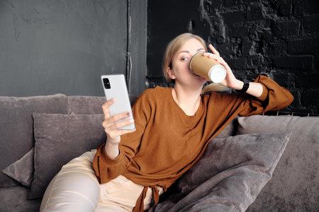 Woman lies on the sofa looks to the phone screen and drinks coffee