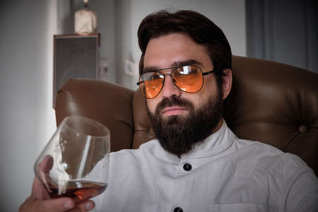 Man dressed in white shirt and yellow eyeglasses holds a glass of cognac and sits in armchair.の写真素材
