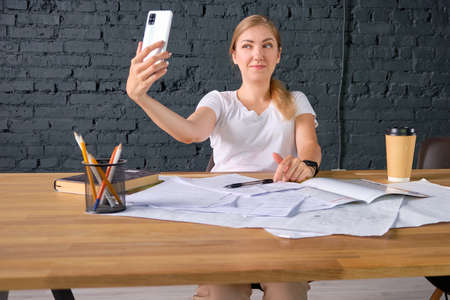 Young woman takes a selfie at a workplace sitting behind work table.