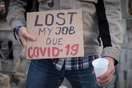 Engineer stands with plastic cup and holds placard Lost my job due covid-19. Construction worker stands opposite construction site with message on poster about unemployment due covid-19 pandemiaの写真素材