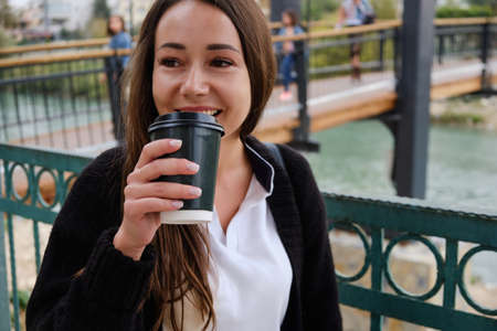 Smiling young woman drinks coffee outside