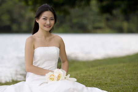 A beautiful young asian woman sits by the edge of a lake in her wedding dressの写真素材