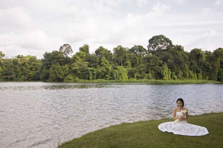 A beautiful asian woman in her wedding dress sits by the side of a lake in Singaporeの写真素材