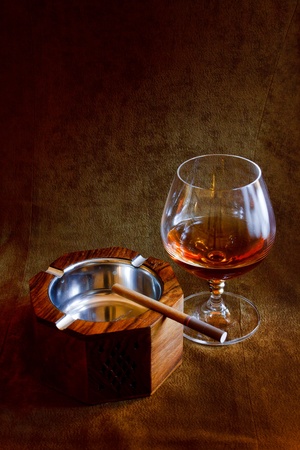 a glass of brandy and a wooden ashtray with a cigarette in the background of brown suedeの写真素材