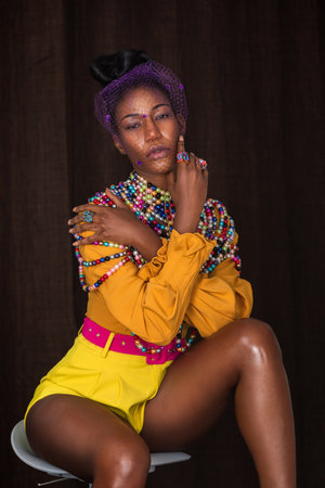 Closeup portrait of a sensual young black woman with long hair, beautiful makeup sitting by herself on a bar chair wearing yellow shorts, pink belt and orange blouse with pearls.の写真素材