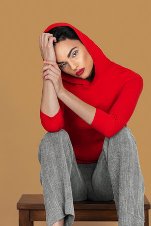 Portrait of a thoughtful young White woman with short black hair, eye makeup and lipstick sitting by herself inside a studio with a pecan background wearing a red hooded sweater.の写真素材