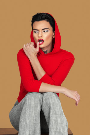Portrait of a sexy young White woman with short black hair, eye makeup and lipstick sitting by herself inside studio with pecan background wearing red hooded sweater and grey pantsの写真素材