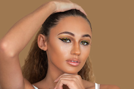 Closeup portrait of a sensual young mixed race woman with long blond hair, light makeup and eye shadow posing by herself touching her head inside a studio with a pecan background.の写真素材