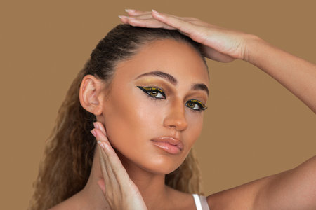 Closeup portrait of a desirable young mixed race woman with long blond hair, light makeup and eye shadow posing by herself touching her head inside a studio with a pecan backgroundの写真素材