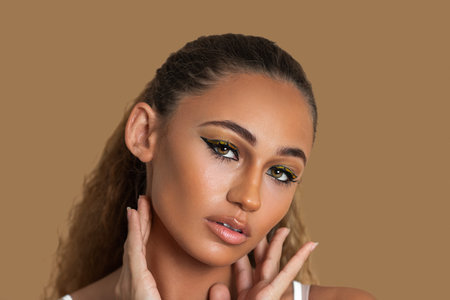 Closeup portrait of a sexy young mixed race woman with long blond hair, light makeup and eye shadow posing by herself touching her head inside a studio with a pecan background.の写真素材
