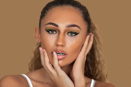 Closeup portrait of a young mixed race woman with long blond hair, light makeup and eye shadow posing by herself touching her head and lips inside a studio with a pecan background.の写真素材