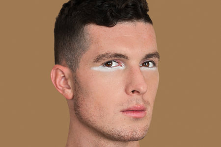 Closeup portrait of a strong young White man with short brown hair, light makeup and white eyeshadow sitting by himself inside studio with pecan background wearing black shirt.の写真素材
