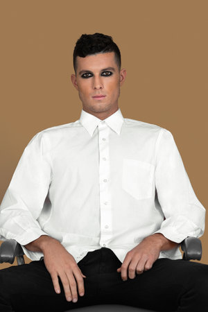 Portrait of a sexy young White man with short brown hair, light makeup and dark eyeshadow sitting by himself inside a studio with a pecan background wearing a white button up shirtの写真素材