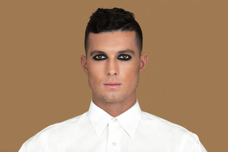 Closeup portrait of handsome young White man with short brown hair, light makeup and dark eyeshadow sitting by himself inside studio with pecan background wearing white shirt.の写真素材