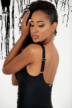 Portrait of a serene young Latin woman with long curly black hair and beautiful makeup standing by herself inside a studio with a black art background wearing a black swimsuit.の写真素材