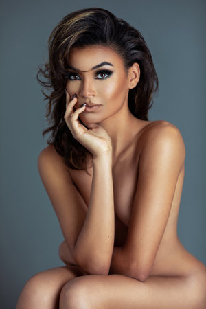 Closeup portrait of a sensual multiracial girl with long wavy brown hair and beautiful makeup sitting naked by herself inside a studio with gray background.の写真素材