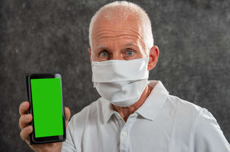 senior man in a medical mask showing smartphone with a green screen. Concept of the danger of coronavirusの写真素材