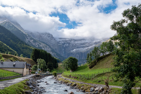 The Cirque of Gavarnie, Pyrenes mountains. The mountains are covered in snow and the sky is cloudy. The river is surrounded by a lush green valleyの写真素材