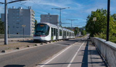 A tramway is traveling down a street. The tram is white and has a green stripe on it. The street is...