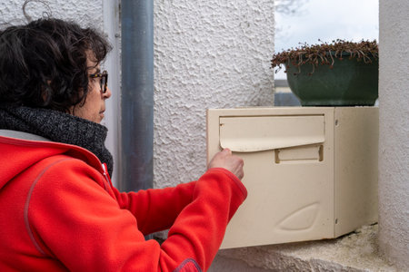 A woman in a red coat opens a mailbox. The box is hanging on the wall.の写真素材